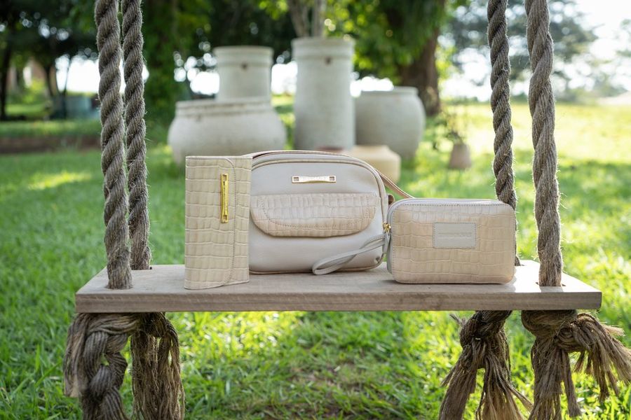 Carteira, bolsa e uma peça menor, todas off-white com textura de croco, lado a lado sobre um balanço de madeira